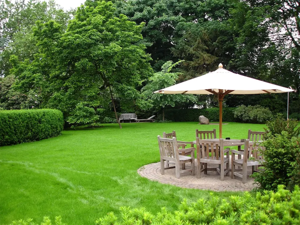 Idyllischer Garten mit üppigen Grünflächen, einem großen Sonnenschirm und einer Holzsitzgruppe auf einem gepflasterten Rundweg, umgeben von dichten Bäumen und Sträuchern.
